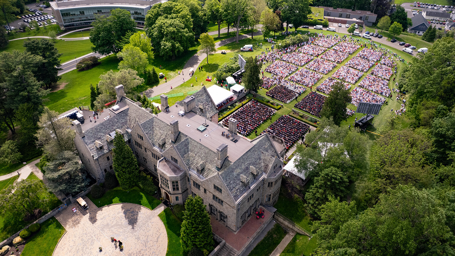 Aerial view of a Bellarmine Hall surrounded by lush trees. Nearby, a crowd gathers on a lawn for an outdoor event, suggesting a celebratory atmosphere.